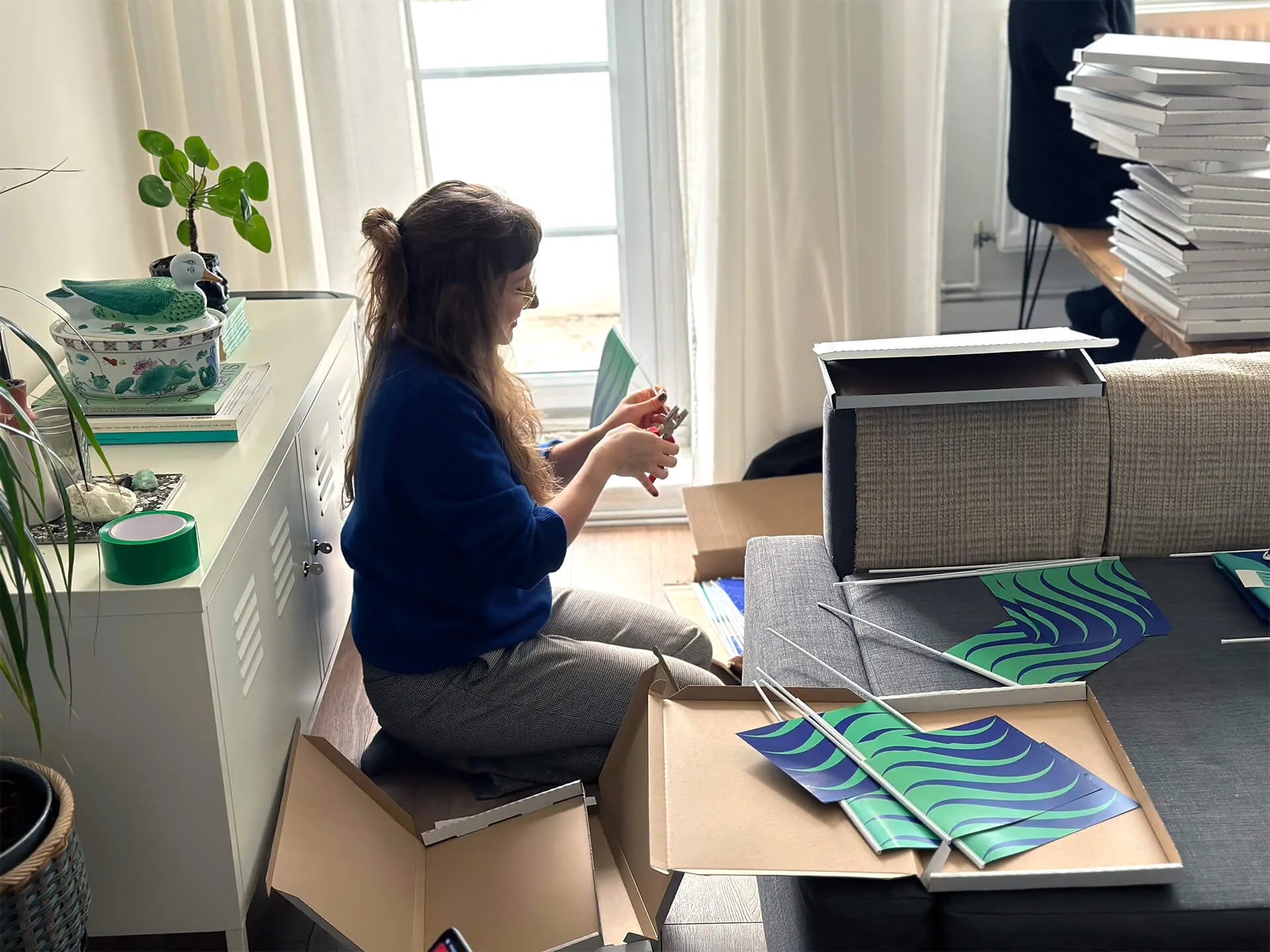 A woman sits on the floor, packing flags into boxes. The flag is seagreen and ultramarine blue waves. 
