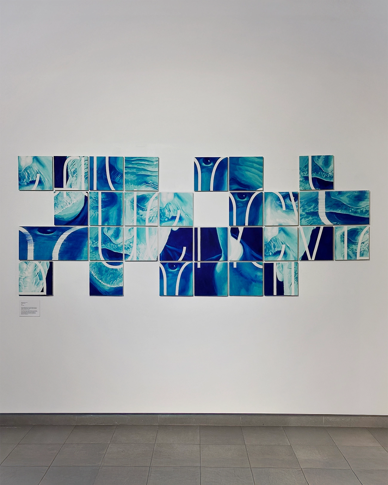 A blue and green painted artwork on a white wall. The artwork is made up of 25 squares, disorganised like a puzzle, showing portraits of two men speaking. 