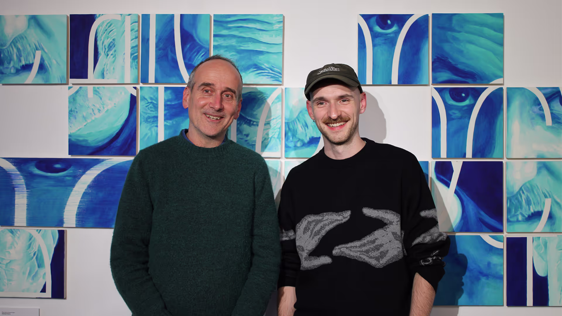 Two men stand in front a blue and green artwork in a gallery.