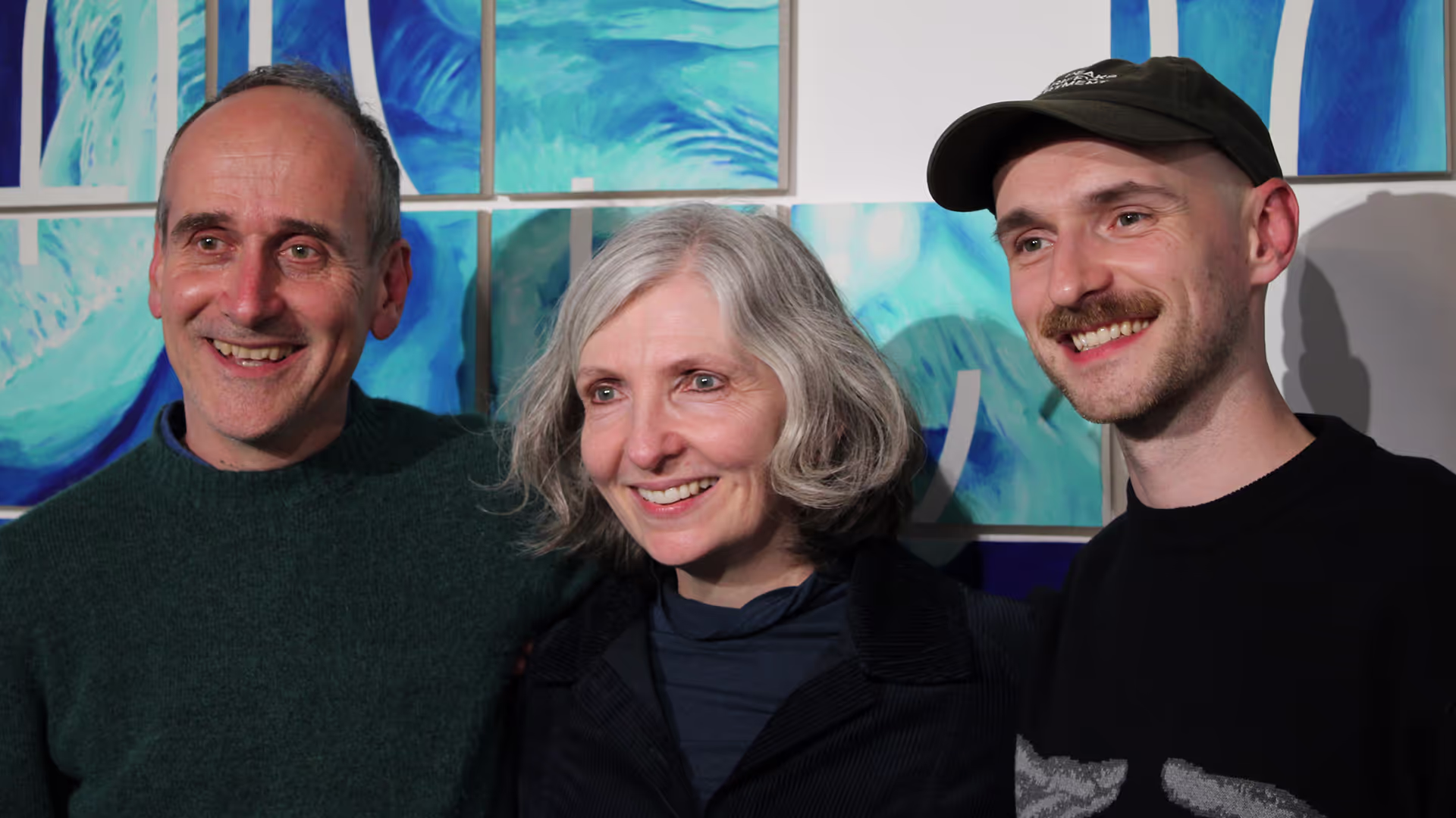 Two men and a woman stand in front a blue and green artwork in a gallery.